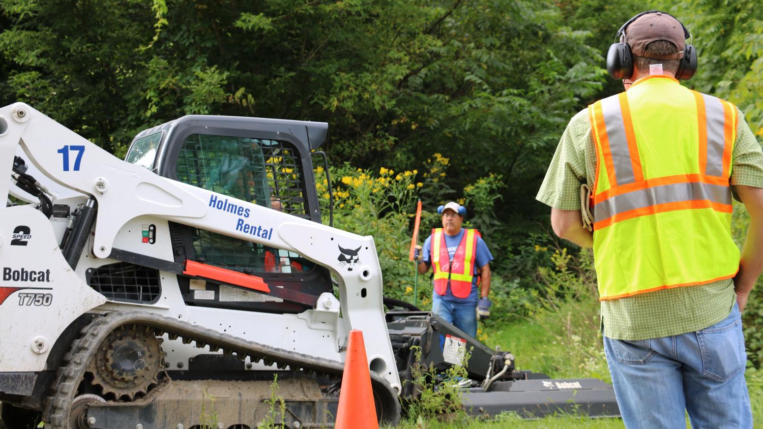 Volunteers wearing protective gear guide a brush trimmer along a future trail.