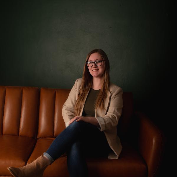 Kayla (she/hers) is seated on a leather couch posing for a photo.