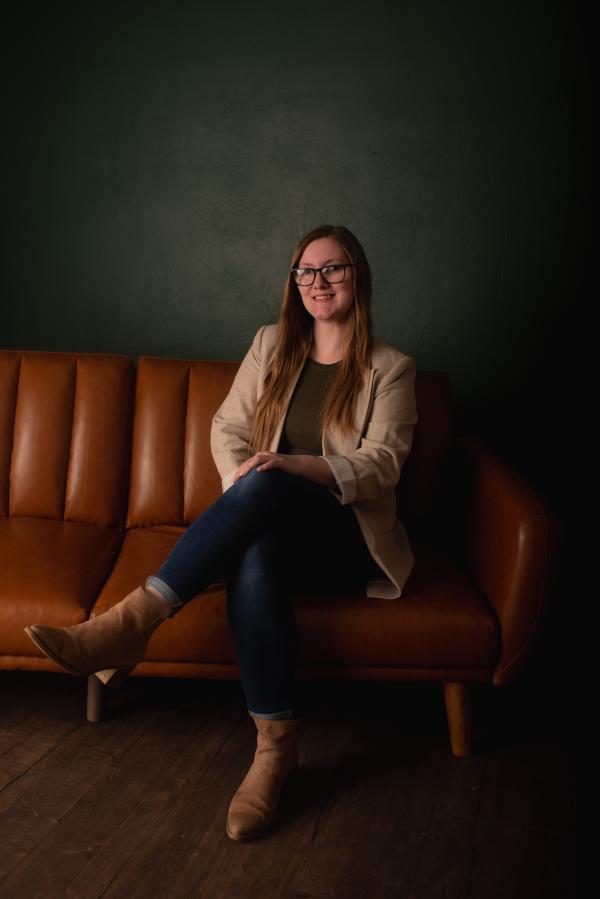 Kayla (she/hers) is seated on a leather couch posing for a photo.